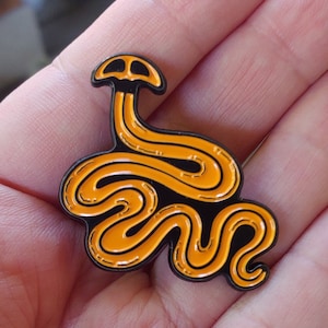 May include: Orange and black enamel pin of a snake with a mushroom head. The snake is curled up and the mushroom head is at the top of the pin.