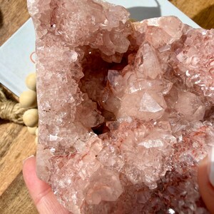 Op de afbeelding: Een grote, ruwe, roze kristalcluster met een ruw, getextureerd oppervlak. De kristal heeft een holle kern en is samengesteld uit vele kleine, individuele kristalpunten. De kleur is zachtroze met enkele gebieden van dieper rood.