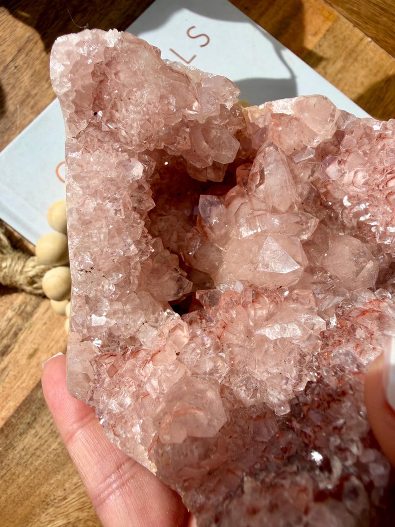 Op de afbeelding: Een grote, ruwe, roze kristalcluster met een ruw, getextureerd oppervlak. De kristal heeft een holle kern en is samengesteld uit vele kleine, individuele kristalpunten. De kleur is zachtroze met enkele gebieden van dieper rood.