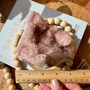 Op de afbeelding: Een grote, roze kristalgeode, ongeveer 7.6 cm breed, ligt op een boek met de titel "Crystals Guide To" met een houten kralenketting. De geode heeft een kristalstructuur en is lichtroze. Het boek is open.