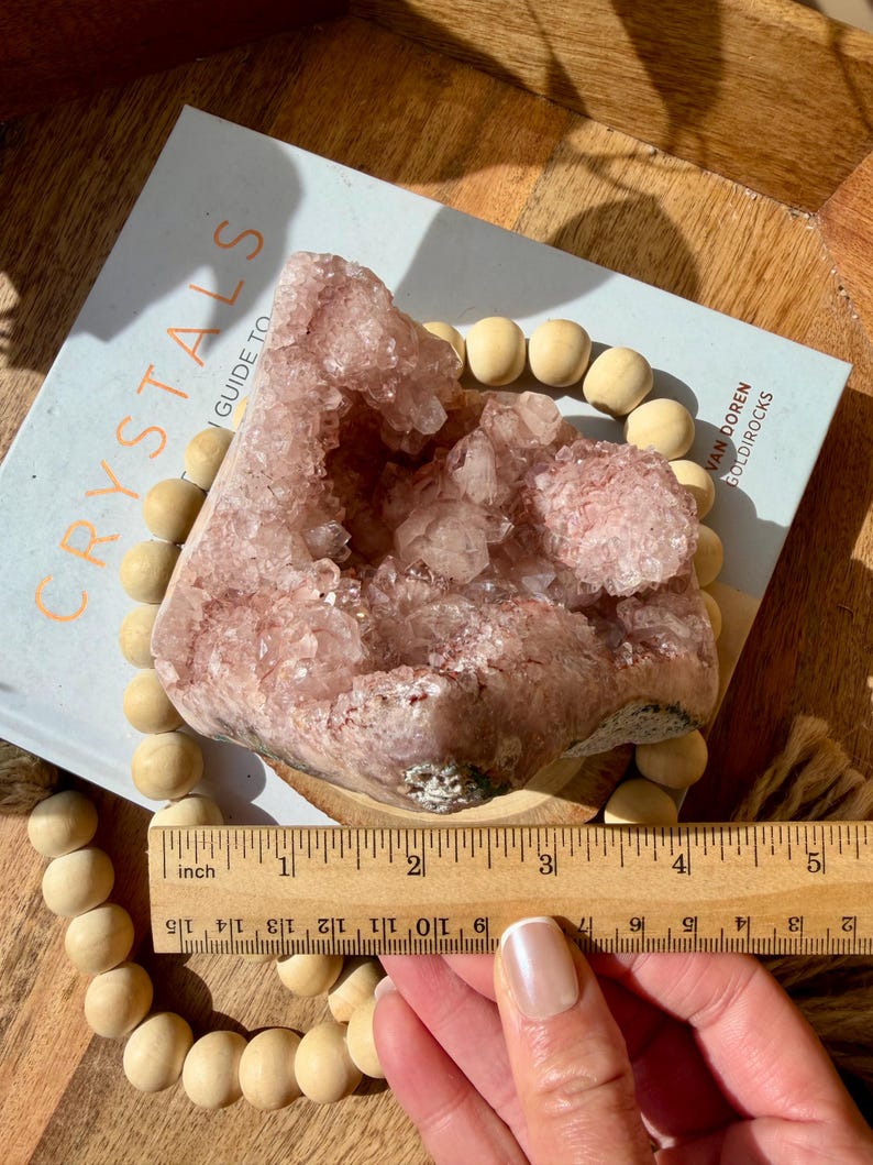 Op de afbeelding: Een grote, roze kristalgeode, ongeveer 7.6 cm breed, ligt op een boek met de titel "Crystals Guide To" met een houten kralenketting. De geode heeft een kristalstructuur en is lichtroze. Het boek is open.