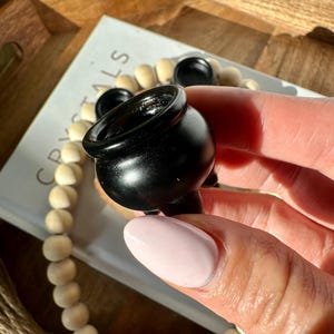 May include: A small, black cauldron with a rounded body and three small feet. The cauldron is held in a hand with a light pink nail polish. The background includes a wooden tray, a beaded garland, and a card with the word "CRYSTALS".
