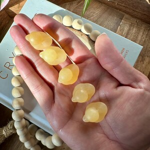 Op de afbeelding: Een hand houdt zes gepolijste, gele calcietstenen vast. De stenen hebben een gladde, afgeronde vorm. Op de achtergrond is een boek met de woorden "CRYSTAL" en "AREN ROCKS" te zien, samen met een houten kralenketting.