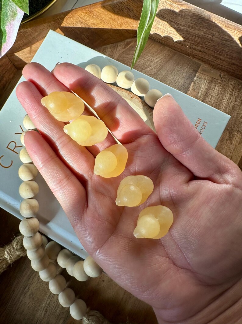 Op de afbeelding: Een hand houdt zes gepolijste, gele calcietstenen vast. De stenen hebben een gladde, afgeronde vorm. Op de achtergrond is een boek met de woorden "CRYSTAL" en "AREN ROCKS" te zien, samen met een houten kralenketting.