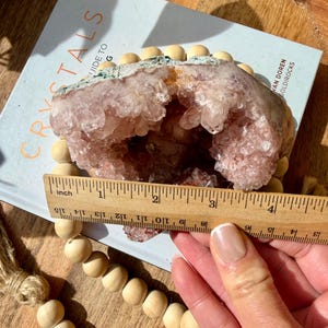 Op de afbeelding: Een lichtroze geodekristal met een hol midden, rustend op een boek met de titel "Crystals Guide To". De geode is ongeveer 10 cm breed. Een houten kralenkrans en een liniaal staan ook op de foto.