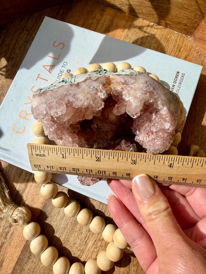 Op de afbeelding: Een lichtroze geodekristal met een hol midden, rustend op een boek met de titel "Crystals Guide To". De geode is ongeveer 10 cm breed. Een houten kralenkrans en een liniaal staan ook op de foto.