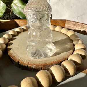 May include: A clear, seated Buddha statue with hands in a prayer position. The statue is on a wooden slice, surrounded by wooden beads. The background includes a plant and a wooden tray, with sunlight casting shadows.