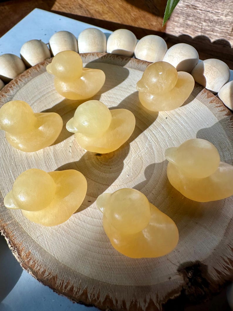 Op de afbeelding: Zes kleine, gele eendenbeeldjes gesneden uit een doorschijnend materiaal. De eenden zijn gerangschikt op een houten schijf, met een natuurlijk houtnerfpatroon. De eenden hebben afgeronde lichamen en kleine, gedetailleerde snavels.