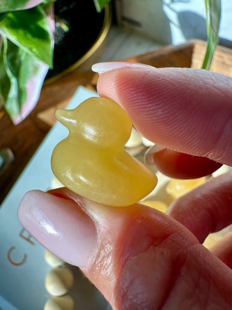 Op de afbeelding: Een kleine, gele eendenfiguur wordt tussen vingers gehouden. De eend heeft een gladde, afgeronde vorm met een kleine snavel en een eenvoudig, charmant ontwerp. De figuur is effen van kleur en lijkt gemaakt van een doorschijnend materiaal.