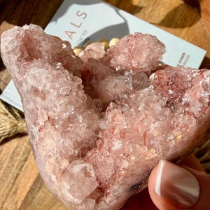 Op de afbeelding: Een grote, roze kristalcluster met een geode-achtige structuur. De kristallen zijn zacht, doorschijnend roze met een sprankelend, getextureerd oppervlak. De kristal wordt in een hand gehouden en een boek met het woord "ALS" is zichtbaar op de achtergrond.