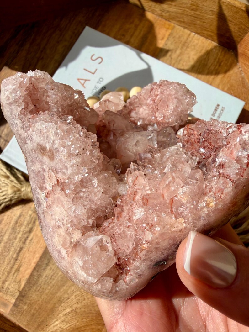 Op de afbeelding: Een grote, roze kristalcluster met een geode-achtige structuur. De kristallen zijn zacht, doorschijnend roze met een sprankelend, getextureerd oppervlak. De kristal wordt in een hand gehouden en een boek met het woord "ALS" is zichtbaar op de achtergrond.