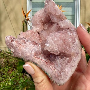 Op de afbeelding: Een grote, ruwe amethistgeode met een lichtpaarse tint. De kristalgroep wordt in een hand gehouden, wat de natuurlijke formatie en kristalstructuur laat zien. De geode heeft een ruwe, getextureerde buitenkant en een sprankelend interieur.