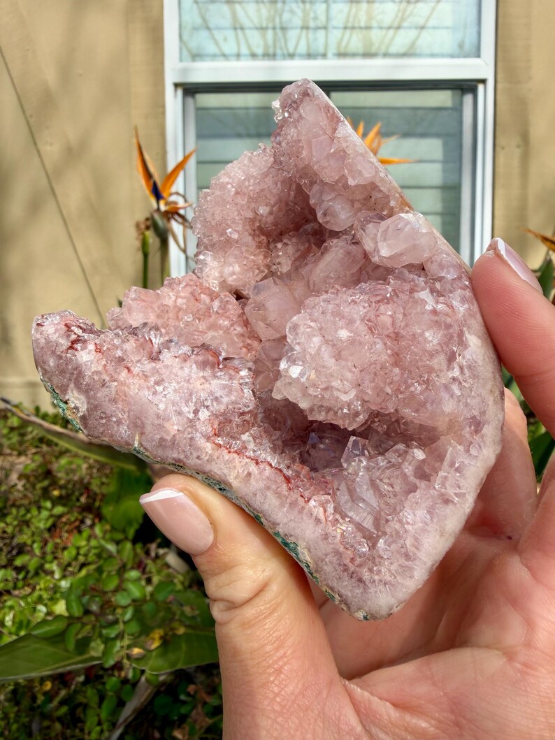 Op de afbeelding: Een grote, ruwe amethistgeode met een lichtpaarse tint. De kristalgroep wordt in een hand gehouden, wat de natuurlijke formatie en kristalstructuur laat zien. De geode heeft een ruwe, getextureerde buitenkant en een sprankelend interieur.