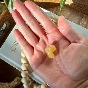 Op de afbeelding: Een kleine, lichtgele kristallen sculptuur in de vorm van een paprika wordt in een open hand gehouden. Het kristal is glad en gepolijst. De achtergrond omvat een houten dienblad, een boek en een streng houten kralen.