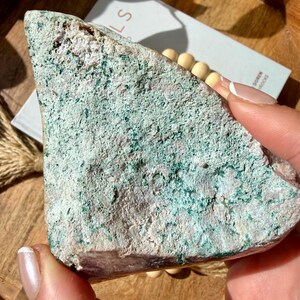 Op de afbeelding: Een ruwe, driehoekige steen met een gevlekte oppervlakte. De steen heeft een mix van lichtroze, witte en turquoise-groene kleuren. De textuur lijkt grof en natuurlijk, wat wijst op een ruwe, ongepolijste staat. De steen wordt in een hand gehouden.