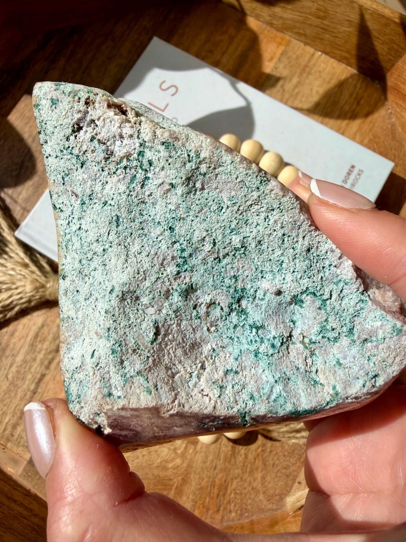 Op de afbeelding: Een ruwe, driehoekige steen met een gevlekte oppervlakte. De steen heeft een mix van lichtroze, witte en turquoise-groene kleuren. De textuur lijkt grof en natuurlijk, wat wijst op een ruwe, ongepolijste staat. De steen wordt in een hand gehouden.