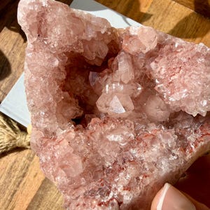 Op de afbeelding: Een grote, ruwe, roze amethistgeode met een kristallijn interieur. De geode heeft een ruwe, getextureerde buitenkant en een holte gevuld met kleinere, sprankelende kristallen. De kleur varieert van lichtroze tot diepere roodachtige tinten.