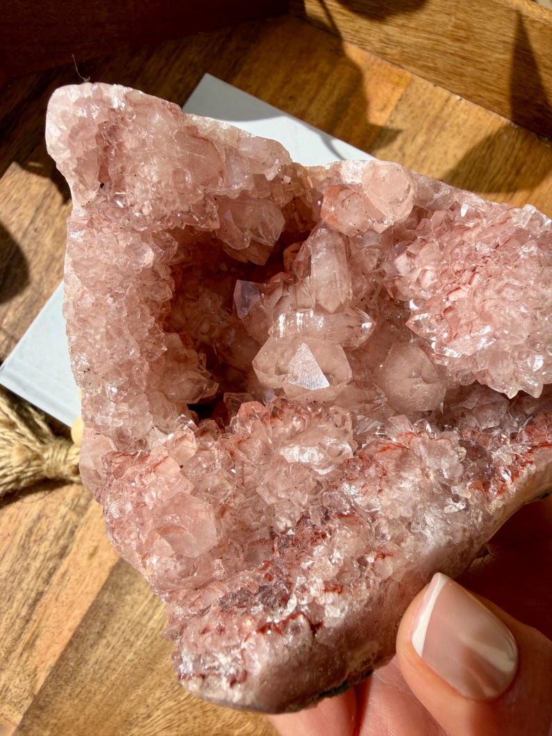 Op de afbeelding: Een grote, ruwe, roze amethistgeode met een kristallijn interieur. De geode heeft een ruwe, getextureerde buitenkant en een holte gevuld met kleinere, sprankelende kristallen. De kleur varieert van lichtroze tot diepere roodachtige tinten.