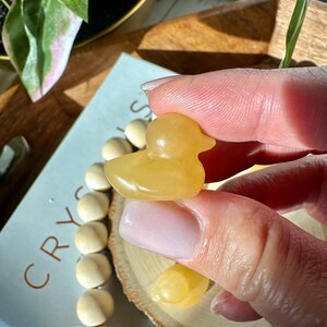 Op de afbeelding: Een kleine, gele eendenfiguur gesneden uit een doorschijnende steen. De eend wordt tussen de vingers gehouden, met een vage achtergrond met een boek met het woord "CRYSTALS" en een houten kralenketting.