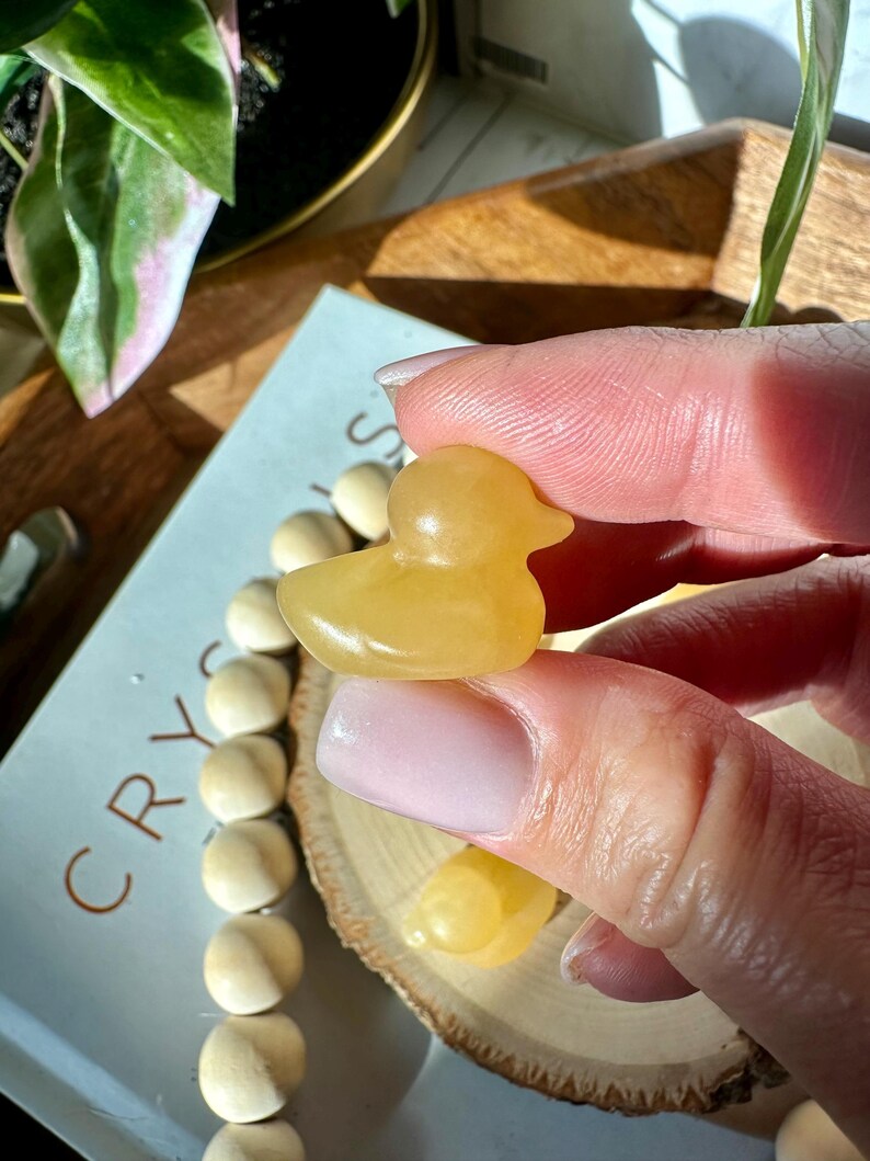 Op de afbeelding: Een kleine, gele eendenfiguur gesneden uit een doorschijnende steen. De eend wordt tussen de vingers gehouden, met een vage achtergrond met een boek met het woord "CRYSTALS" en een houten kralenketting.
