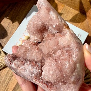 Op de afbeelding: Een grote, roze kristalcluster met een ruw, getextureerd oppervlak. Het kristal heeft een geode-achtig uiterlijk, met een hol centrum gevuld met kleinere, sprankelende kristallen. De kleur is een zacht, stoffig roze met hints van rood en bruin.