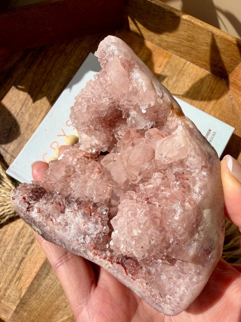 Op de afbeelding: Een grote, roze kristalcluster met een ruw, getextureerd oppervlak. Het kristal heeft een geode-achtig uiterlijk, met een hol centrum gevuld met kleinere, sprankelende kristallen. De kleur is een zacht, stoffig roze met hints van rood en bruin.