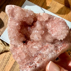 Op de afbeelding: Een grote, roze kristallen geode met een ruwe, getextureerde buitenkant en een cluster van kleinere kristallen binnenin. De geode heeft een natuurlijke, organische vorm.