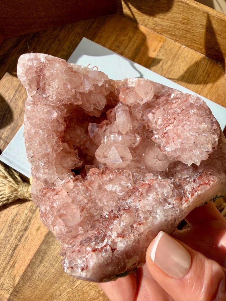 Op de afbeelding: Een grote, roze kristallen geode met een ruwe, getextureerde buitenkant en een cluster van kleinere kristallen binnenin. De geode heeft een natuurlijke, organische vorm.
