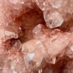 Op de afbeelding: Close-up van een cluster van roze kwartskristallen. De kristallen zijn doorschijnend en hebben scherpe, puntige randen. De algehele textuur is ruw en kristallijn, met variaties in kleur en lichtreflectie.