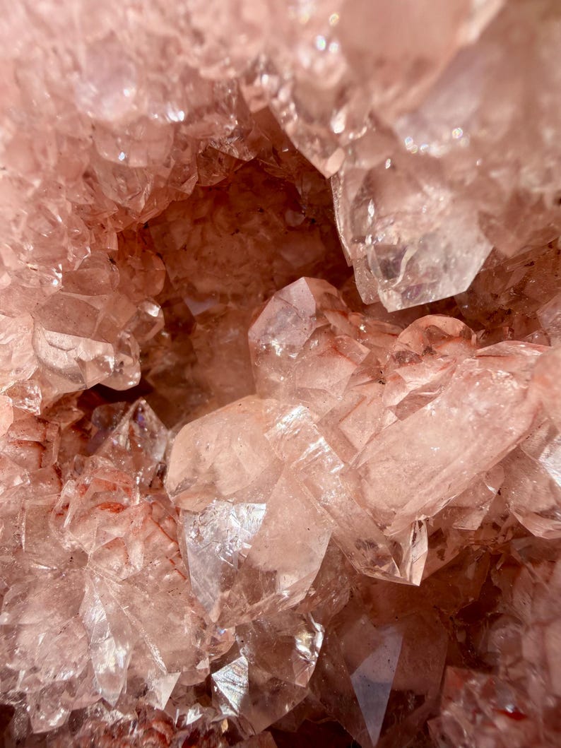 Op de afbeelding: Close-up van een cluster van roze kwartskristallen. De kristallen zijn doorschijnend en hebben scherpe, puntige randen. De algehele textuur is ruw en kristallijn, met variaties in kleur en lichtreflectie.