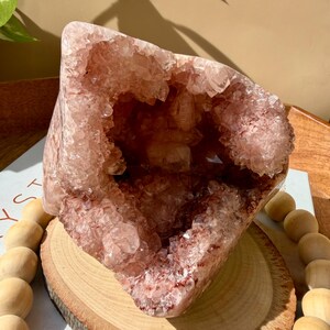 Op de afbeelding: Een grote, roze amethist geode kristal met een holle kern, gepresenteerd op een houten schijf. De geode heeft een ruwe, kristallijne buitenkant en een diepe, open holte. De geode is omgeven door houten kralen.