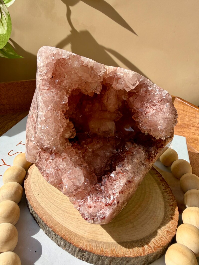 Op de afbeelding: Een grote, roze amethist geode kristal met een holle kern, gepresenteerd op een houten schijf. De geode heeft een ruwe, kristallijne buitenkant en een diepe, open holte. De geode is omgeven door houten kralen.