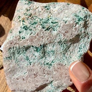 Op de afbeelding: Close-up van een natuursteen met een ruwe textuur. De steen heeft een mix van lichtroze, wit en groene vlekken. De steen wordt door een hand vastgehouden.