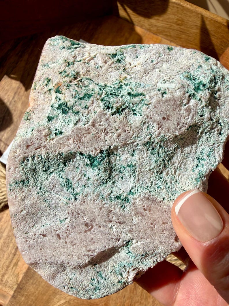 Op de afbeelding: Close-up van een natuursteen met een ruwe textuur. De steen heeft een mix van lichtroze, wit en groene vlekken. De steen wordt door een hand vastgehouden.