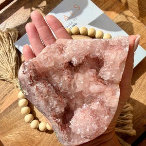 Op de afbeelding: Een grote, roze kristallen geode met een ruw, getextureerd oppervlak. De geode wordt in een hand gehouden en onthult de ingewikkelde kristalstructuur en verschillende tinten roze en rood. Een boek met de titel "Guide to Healing" is zichtbaar.