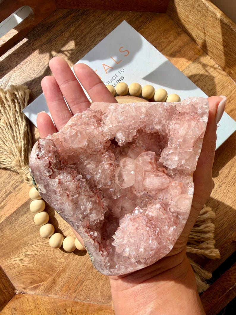 Op de afbeelding: Een grote, roze kristallen geode met een ruw, getextureerd oppervlak. De geode wordt in een hand gehouden en onthult de ingewikkelde kristalstructuur en verschillende tinten roze en rood. Een boek met de titel "Guide to Healing" is zichtbaar.