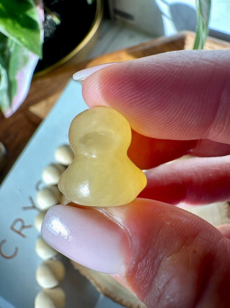 Op de afbeelding: Een kleine, geel getinte kristallen beeldje dat tussen vingers wordt gehouden. Het beeldje heeft een afgerond lichaam en een kopachtige vorm. De achtergrond omvat een lichtblauw oppervlak met het woord "CRY" en een reeks witte kralen.