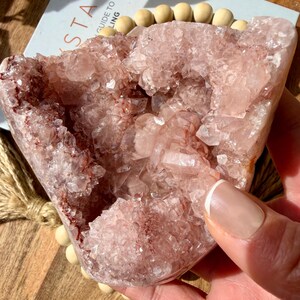 Op de afbeelding: Een grote, roze kristalgeode met een ruw, getextureerd oppervlak. De geode heeft een lichtroze kleur met donkerdere rode en bruine insluitsels. De geode wordt in een hand gehouden. Op de achtergrond staat een boek met het woord "Crystals".