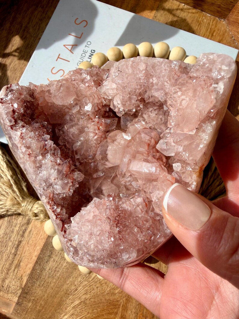 Op de afbeelding: Een grote, roze kristalgeode met een ruw, getextureerd oppervlak. De geode heeft een lichtroze kleur met donkerdere rode en bruine insluitsels. De geode wordt in een hand gehouden. Op de achtergrond staat een boek met het woord "Crystals".