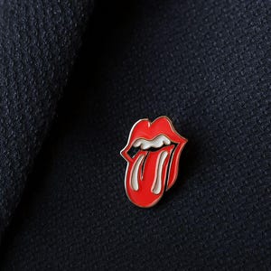 Puede incluir: Un pin de esmalte rojo y blanco con forma de boca con una lengua que sobresale. El pin tiene un borde dorado y está colocado sobre un fondo de tela azul oscuro. Un accesorio popular con temática musical.