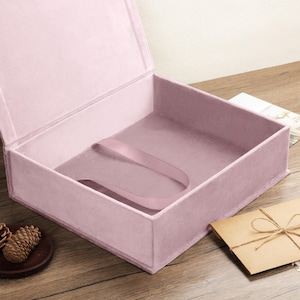 May include: A square, open storage box covered in soft, light pink velvet fabric. The interior features a matching light pink velvet lining and a single, wide, light pink satin ribbon loop attached to the base for lifting or securing items.