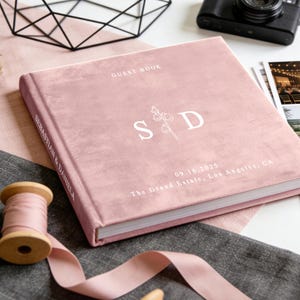 May include: A pink velvet guest book with white text. The cover reads "GUEST BOOK" and features the initials "S D" with a floral design. The book is personalized with names and a wedding date.