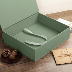 May include: A sage green rectangular gift box with a hinged lid and a light green ribbon attached to the interior base. The box is open, revealing a smooth, solid-colored interior surface, and it features a clean, structured design.
