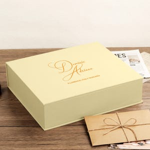 May include: A rectangular, pale yellow keepsake box with a hinged lid. The top features gold-colored script text that reads "Dominic Adriana" and "FLORENCE, ITALY 10.03.2026". The box has a smooth, matte finish and a clean, structured design.