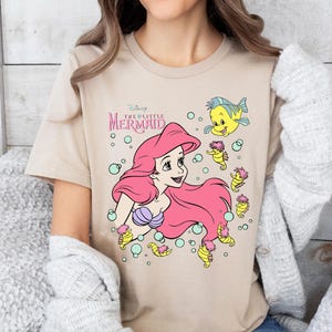 Retro vintage Ariel The Little Mermaid shirt featuring a nostalgic design with Ariel and Flounder. Made from soft, comfortable fabric for all-day wear. Perfect for Disney fans, princess lovers, and classic animation enthusiasts. Great for theme park visits, casual outfits, or as a unique gift. Available in multiple sizes and colors to match any style. A stylish and fun way to showcase love for this beloved Disney character.