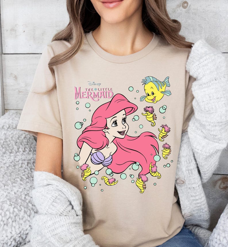 Retro vintage Ariel The Little Mermaid shirt featuring a nostalgic design with Ariel and Flounder. Made from soft, comfortable fabric for all-day wear. Perfect for Disney fans, princess lovers, and classic animation enthusiasts. Great for theme park visits, casual outfits, or as a unique gift. Available in multiple sizes and colors to match any style. A stylish and fun way to showcase love for this beloved Disney character.