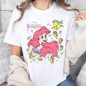 Retro vintage Ariel The Little Mermaid shirt featuring a nostalgic design with Ariel and Flounder. Made from soft, comfortable fabric for all-day wear. Perfect for Disney fans, princess lovers, and classic animation enthusiasts. Great for theme park visits, casual outfits, or as a unique gift. Available in multiple sizes and colors to match any style. A stylish and fun way to showcase love for this beloved Disney character.