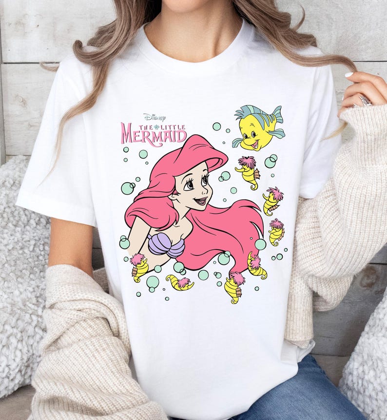 Retro vintage Ariel The Little Mermaid shirt featuring a nostalgic design with Ariel and Flounder. Made from soft, comfortable fabric for all-day wear. Perfect for Disney fans, princess lovers, and classic animation enthusiasts. Great for theme park visits, casual outfits, or as a unique gift. Available in multiple sizes and colors to match any style. A stylish and fun way to showcase love for this beloved Disney character.