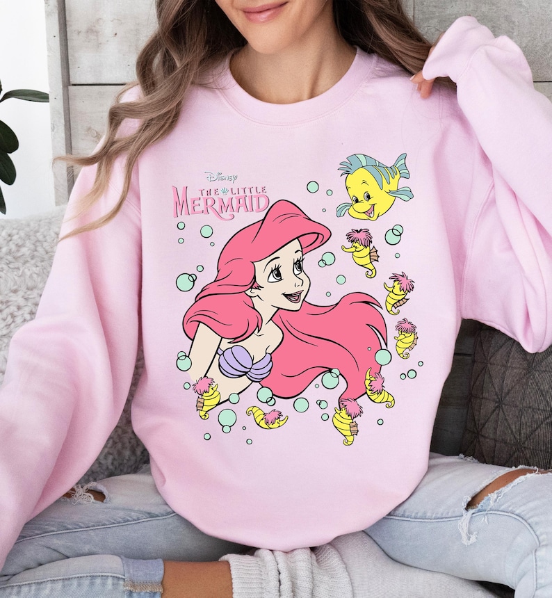 Retro vintage Ariel The Little Mermaid shirt featuring a nostalgic design with Ariel and Flounder. Made from soft, comfortable fabric for all-day wear. Perfect for Disney fans, princess lovers, and classic animation enthusiasts. Great for theme park visits, casual outfits, or as a unique gift. Available in multiple sizes and colors to match any style. A stylish and fun way to showcase love for this beloved Disney character.