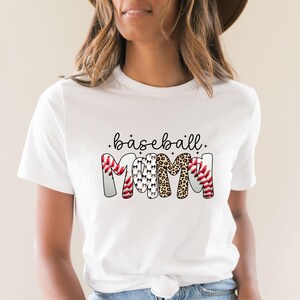 Peut inclure: T-shirt blanc avec l'inscription "baseball mom" en &eacute;criture noire. Le mot "MOM" est con&ccedil;u avec des balles de baseball, des silhouettes de chiens et des motifs l&eacute;opard. Un t-shirt d&eacute;contract&eacute; pour tous les jours.